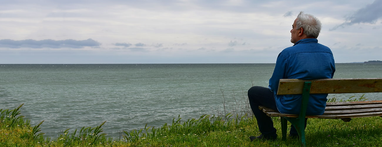 Senior man watching the horizon sitting on a bench by the see in a dark cloudy day
