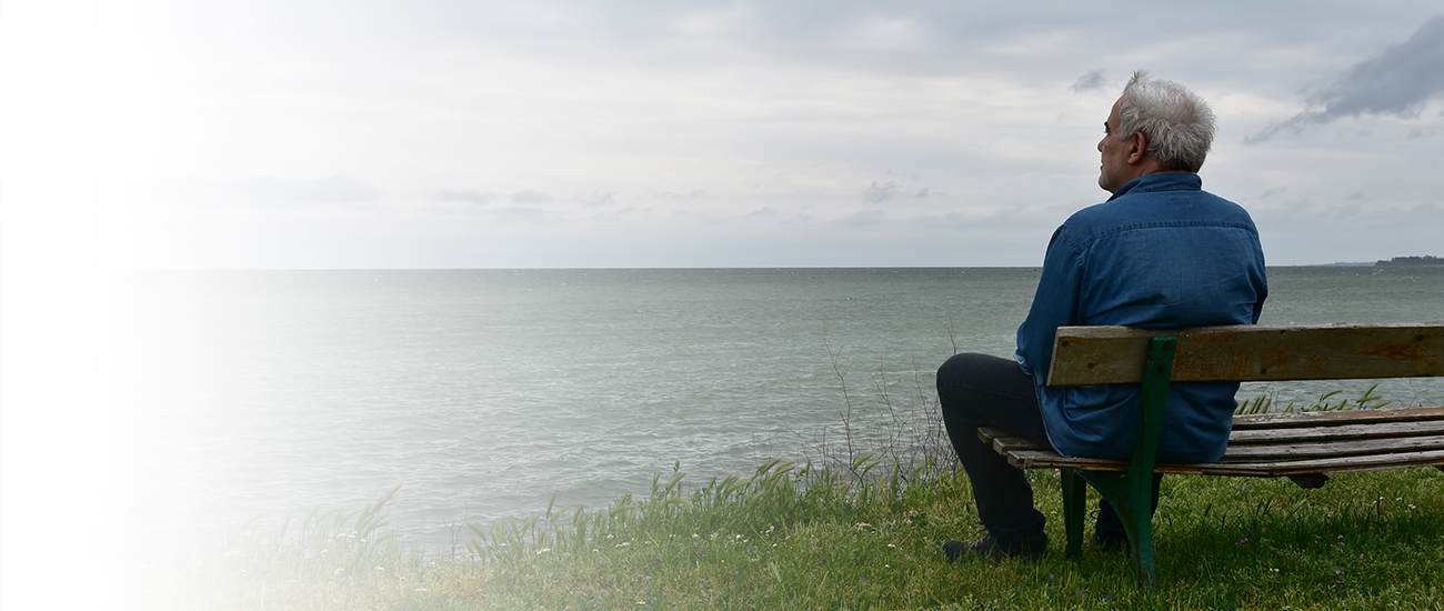 Senior man watching the horizon sitting on a bench by the see in a dark cloudy day