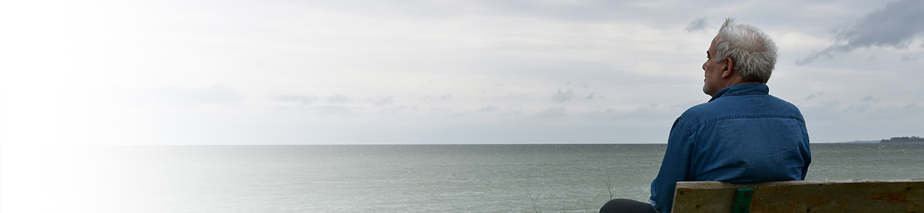 Senior man watching the horizon sitting on a bench by the see in a dark cloudy day
