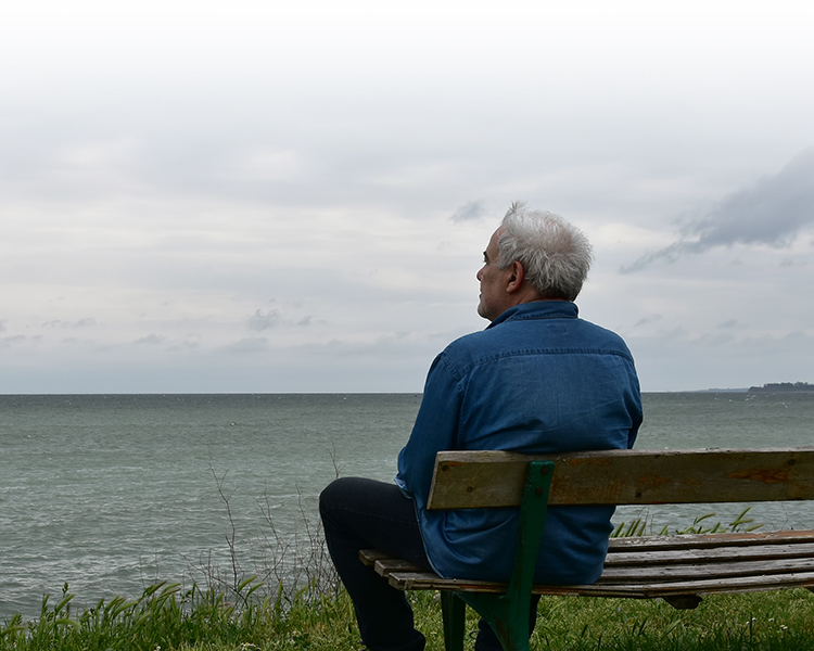 Senior man watching the horizon sitting on a bench by the see in a dark cloudy day